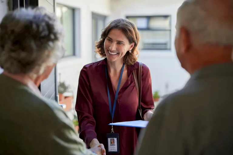 A smiling professional woman in a burgundy blouse and blue lanyard shaking hands with an elderly couple at the entrance of their modern home.
